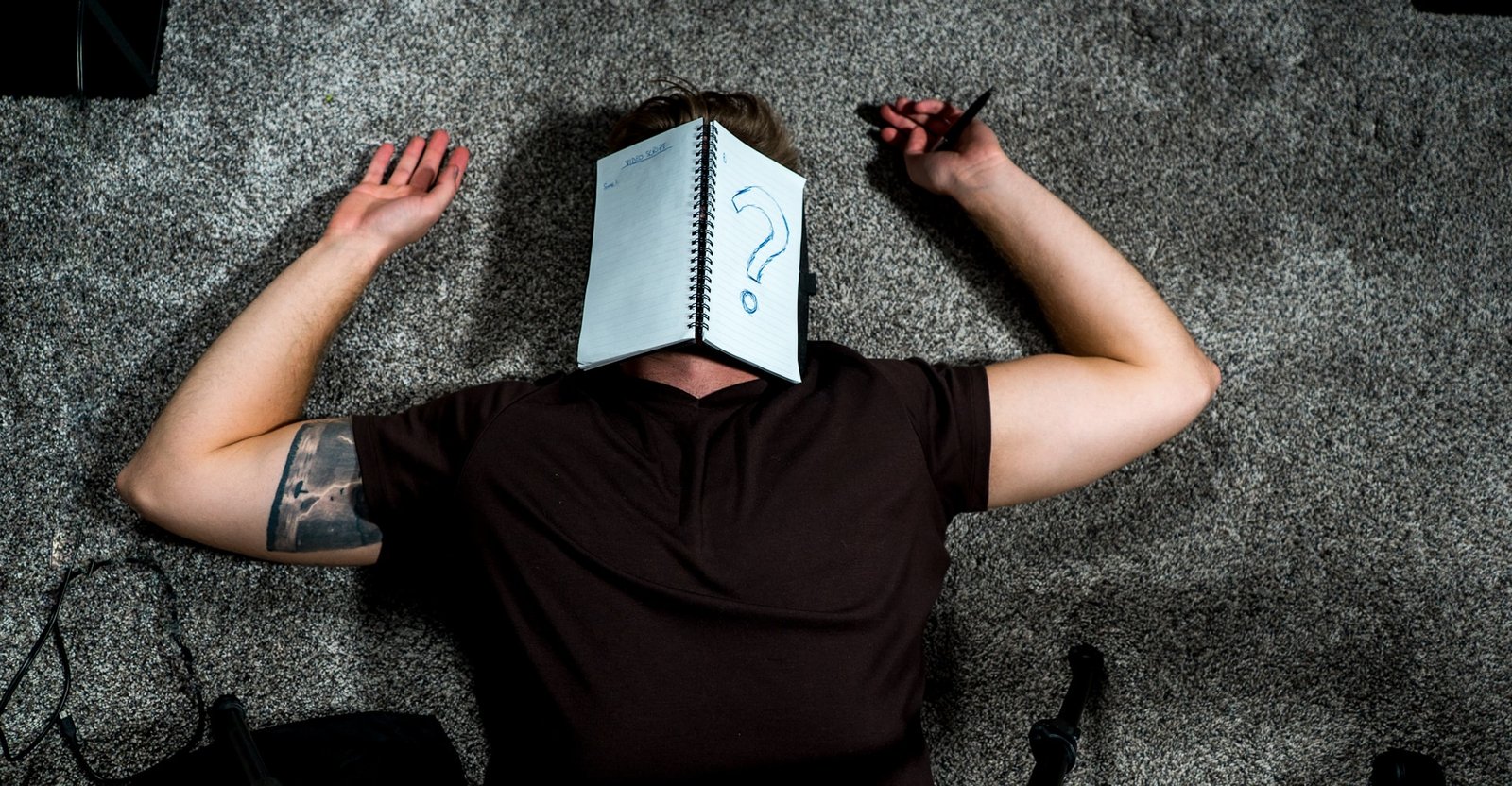 Person lying on the floor with a notebook covering their face, a large question mark drawn on the page — a humorous take on writer’s block for the CMeewrites About Me page.