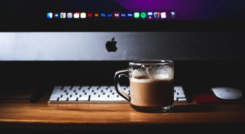Glass mug of coffee on a wooden desk in front of an Apple iMac — a minimalist version of the CMeewrites writing setup, featured on the About Me page.